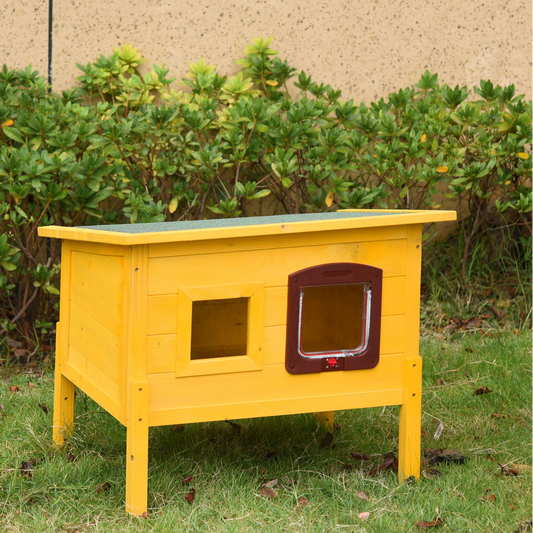 Yellow pet house with a brown door on a grassy area with green bushes in the background