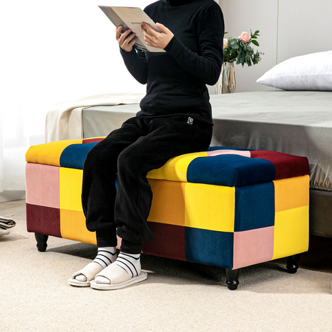 Person sitting on a colorful patchwork ottoman reading a book in a bedroom.