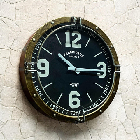 Round wall clock with black face and brass frame on a textured beige wall.