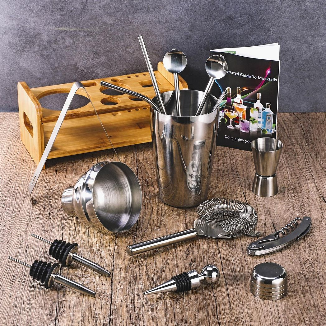 Set of bar tools including a cocktail shaker, jigger, and other utensils on a wooden surface with a background of bottles and a book.
