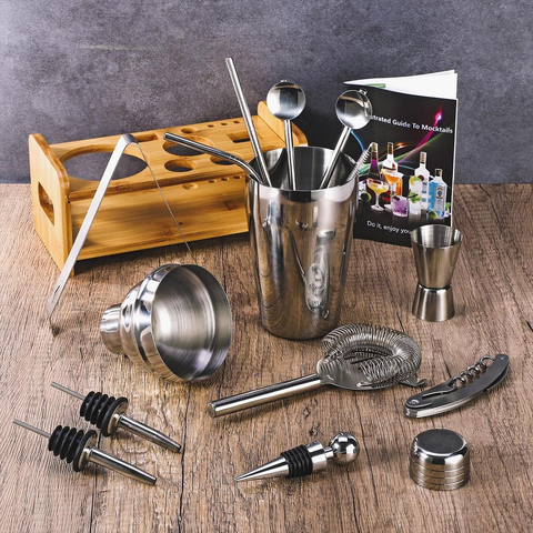 Set of bar tools including a cocktail shaker, jigger, and other utensils on a wooden surface with a background of bottles and a book.