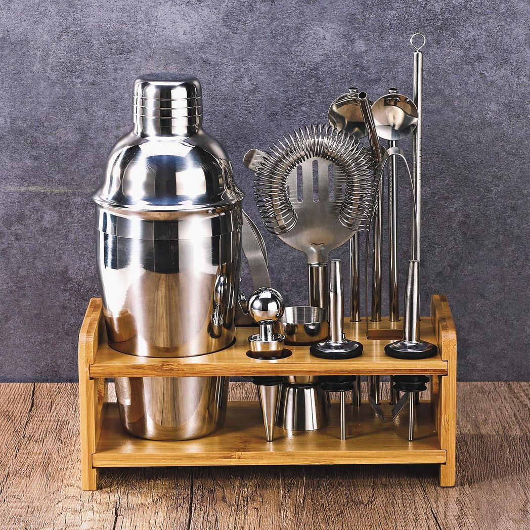 Set of cocktail-making tools including a shaker and strainers on a wooden stand against a gray background.