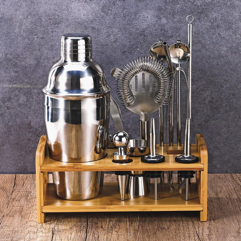 Set of cocktail-making tools including a shaker and strainers on a wooden stand against a gray background.